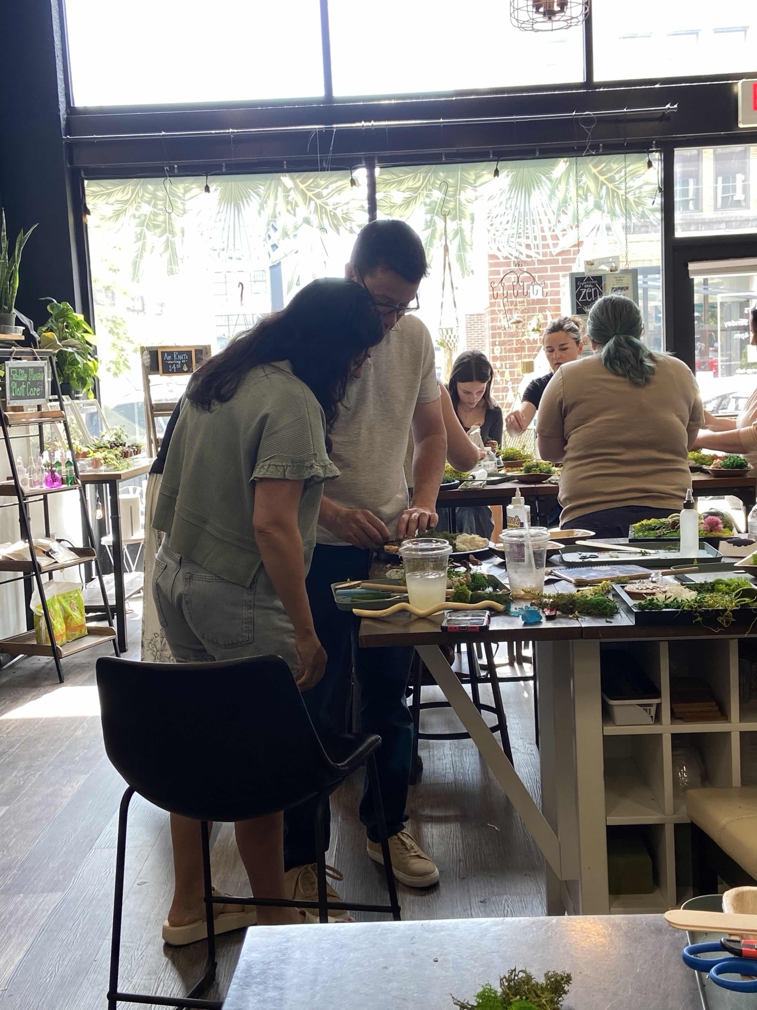 A room of guests standing and seated at the long table building terrariums, storefront windows behind them