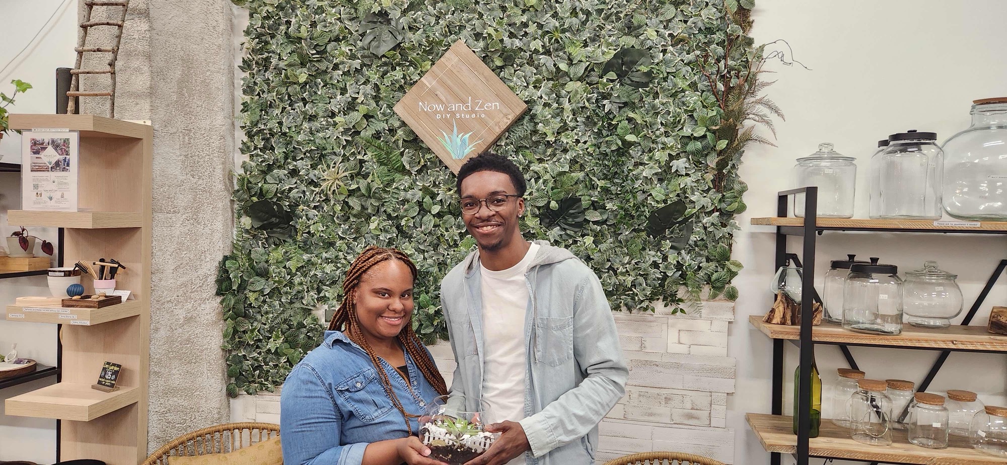 Two friends smiling in front of the living plant wall and Now and Zen sign, holding a finished terrarium