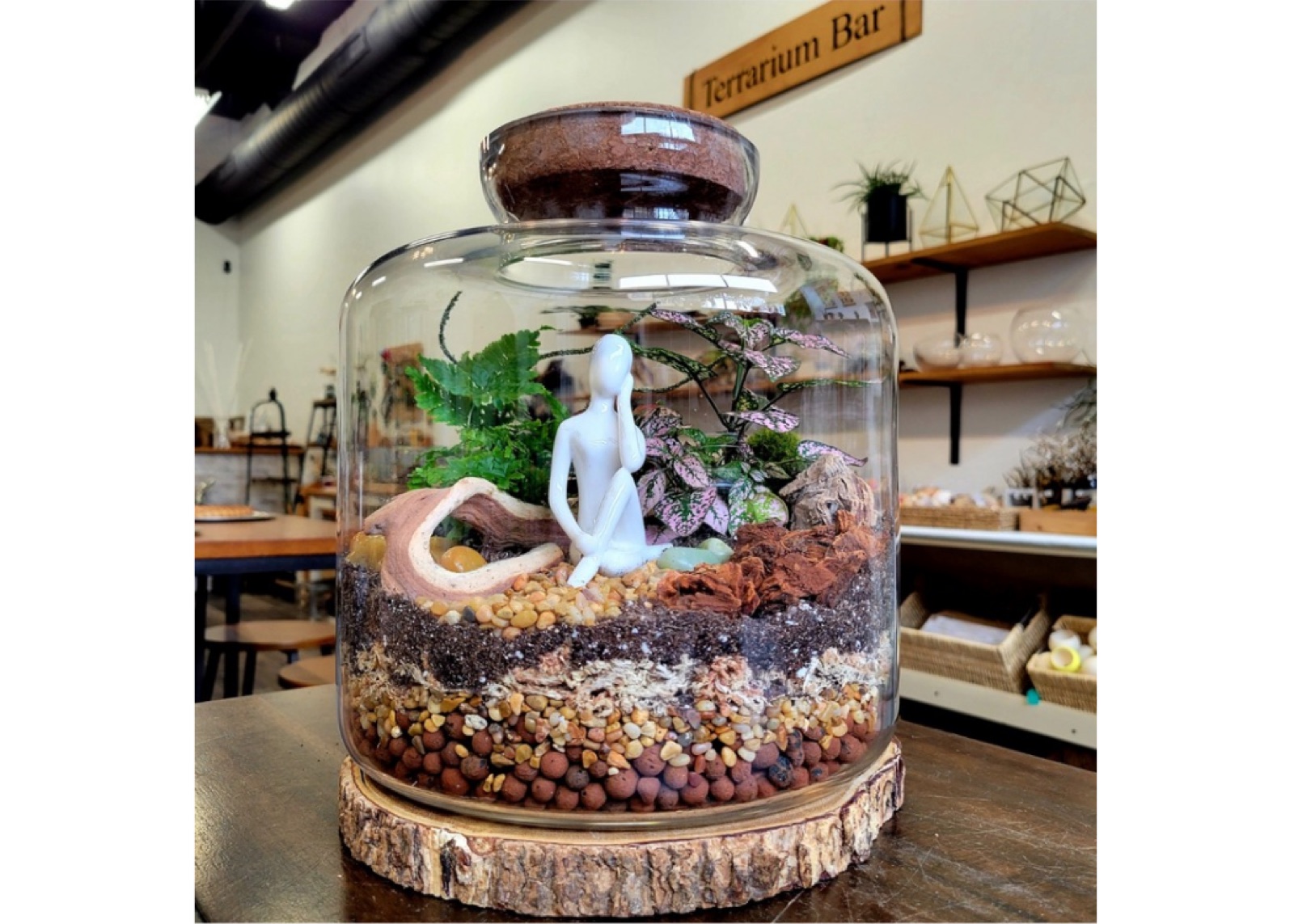 A tall cloche terrarium with a meditating figure and layered stones, on a wood slice under the Terrarium Bar sign