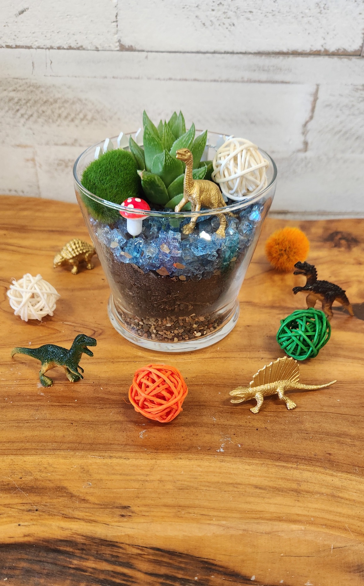 Dinosaur Fun, a glass terrarium with a succulent, blue stones, and toy dinosaurs staged around it