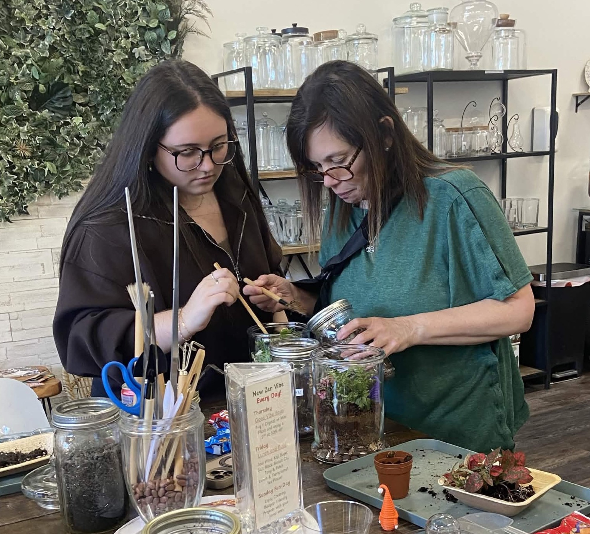 Two guests building terrariums side by side at the studio bar, shelves of glass vessels behind them