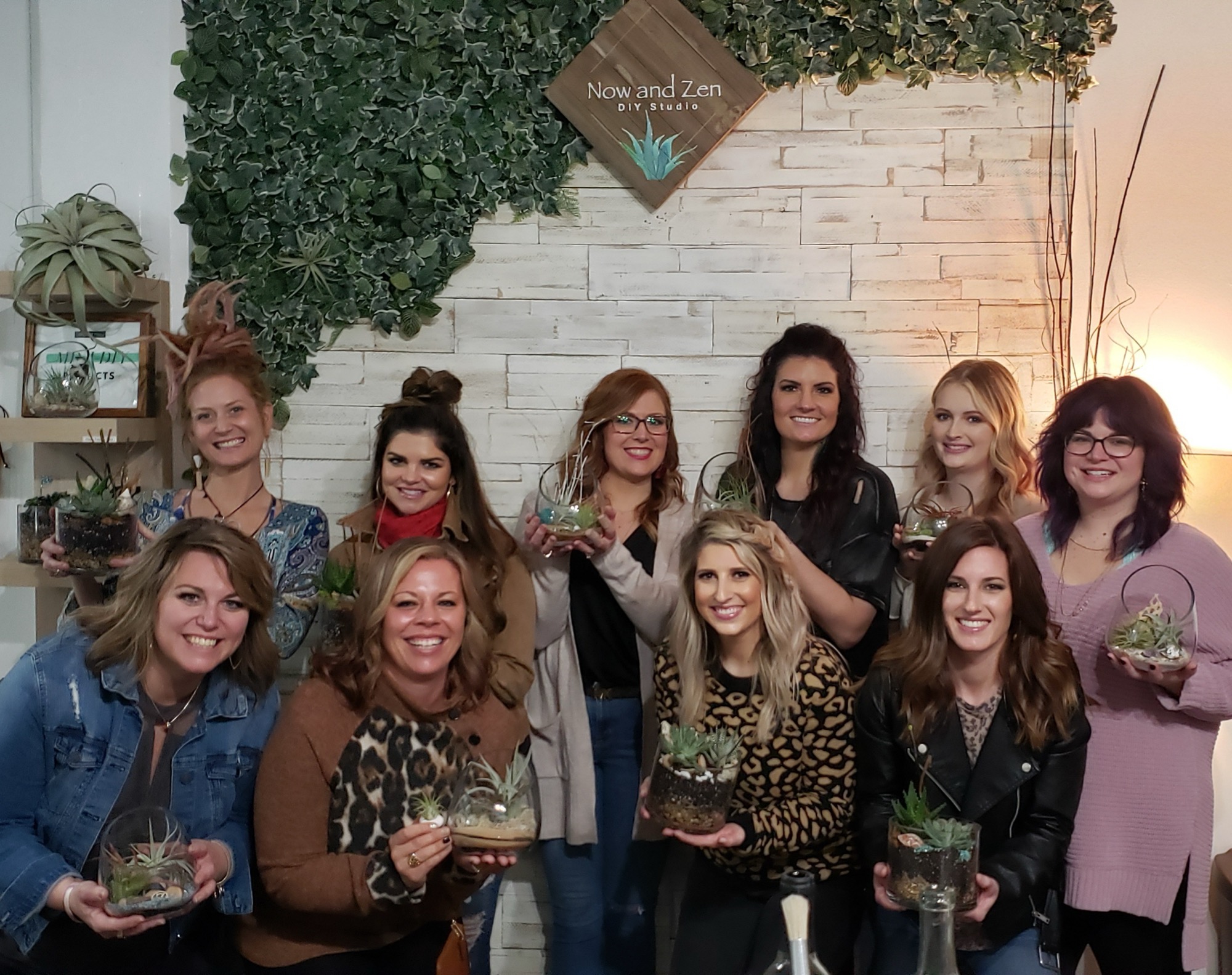 A group of guests holding up the terrariums they built together, in front of the plant wall and Now and Zen sign