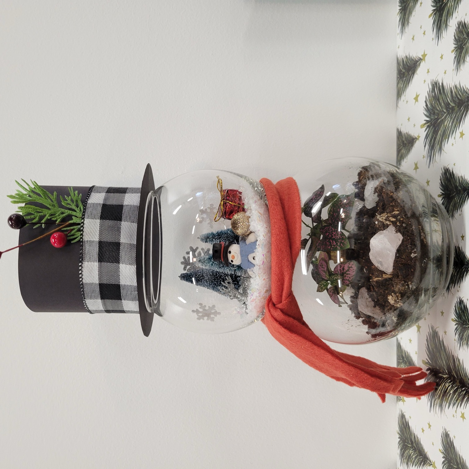 A snowman built from two stacked glass globes, a top hat and red scarf, with a winter scene above and planted foliage below
