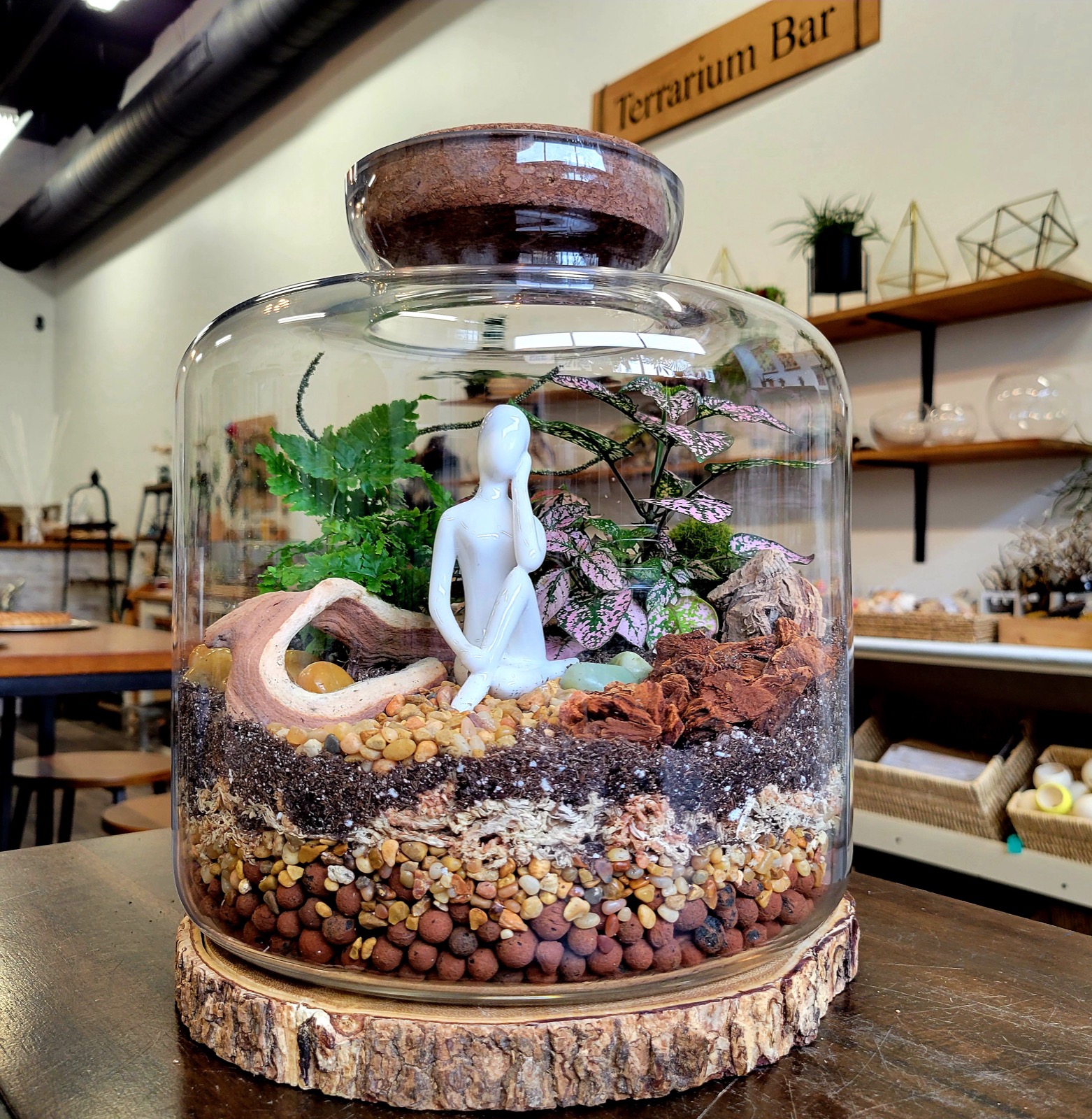 A large closed cloche terrarium with ferns, pink polka dot plants, driftwood and a seated figure over layered stone and soil, under the Terrarium Bar sign