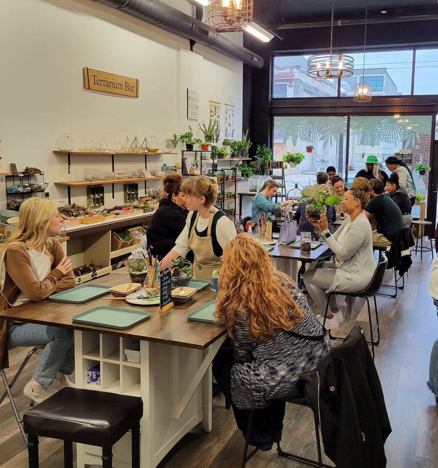 A busy afternoon at the Terrarium Bar, guests seated at tables building terrariums under the Terrarium Bar sign with the storefront windows behind them