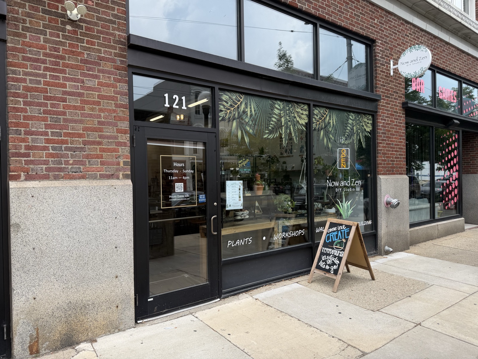 The Now and Zen storefront from the sidewalk, blade sign and a come-in-and-create-terrariums sandwich board out front